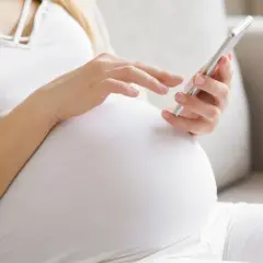 Kobieta w 24. tygodniu ciąży (l4 w ciąży) przegląda coś na telefonie, siedząc na kanapie.