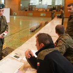 Młody rezerwista w zielonym beretku trzyma dokumenty. W tle inni żołnierze i sala gimnastyczna.