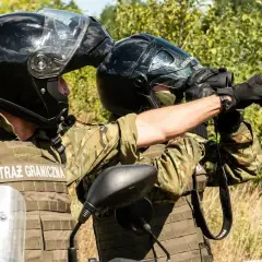 Strażnik graniczny w kamizelce taktycznej i kasku obserwuje teren przez lornetkę, wskazując kierunek.