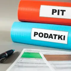 Dwa segregator, jeden czerwony z napisem PIT, drugi niebieski z napisem PODATKI, leżą na biurku obok formularzy podatkowych i długopisu. Czas na zwolnienie z PIT!