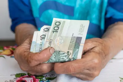 Dłonie starszej osoby trzymają banknoty 100 zł. Czy to wystarczy na godne życie przy minimalnej emeryturze w Polsce?