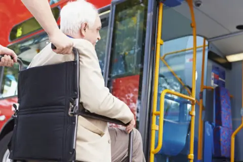 Starszy pan na wózku inwalidzkim, prawdopodobnie z chorobami kwalifikującymi do **2 grupa inwalidzka**, wsiada do autobusu z pomocą.