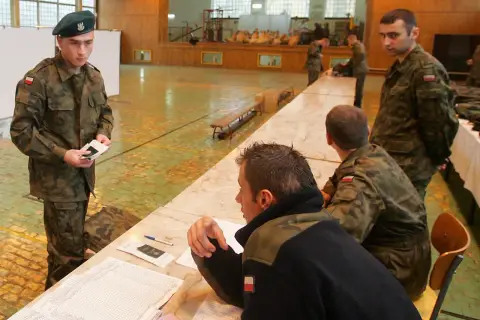 Młody rezerwista w zielonym beretku trzyma dokumenty. W tle inni żołnierze i sala gimnastyczna.