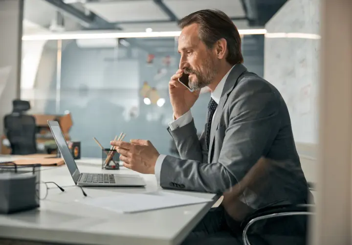 Mężczyzna w garniturze rozmawia przez telefon, patrząc na laptopa. To typowy dzień pracy CEO, co to znaczy? To znaczy, że zarządza firmą.