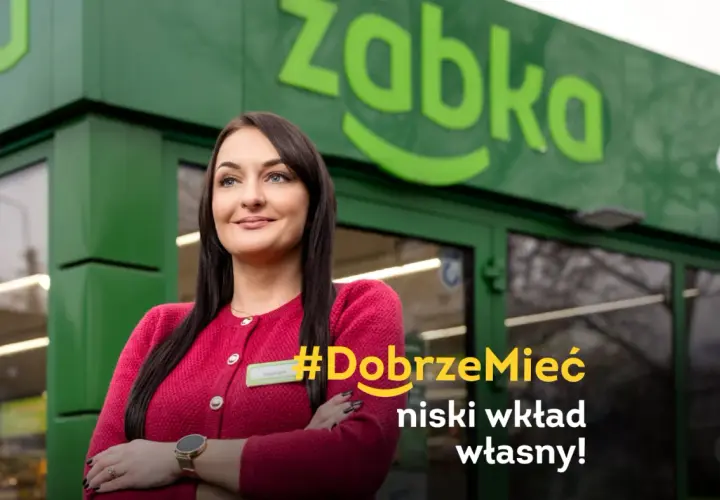 Uśmiechnięta franczyzobiorczyni Żabki przed sklepem. Hasło: #DobrzeMieć niski wkład własny!