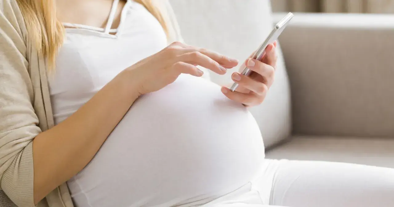 Kobieta w 24. tygodniu ciąży (l4 w ciąży) przegląda coś na telefonie, siedząc na kanapie.