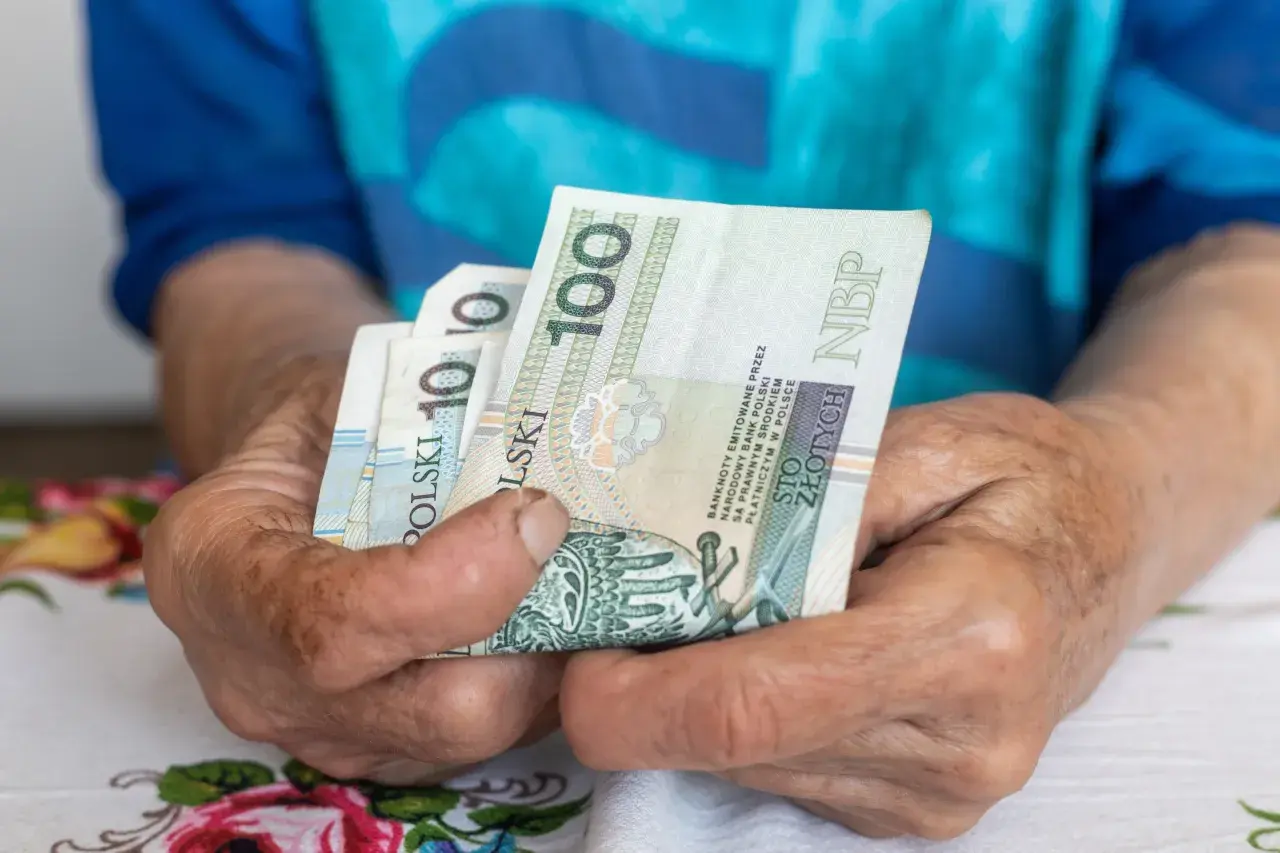 Dłonie starszej osoby trzymają banknoty 100 zł. Czy to wystarczy na godne życie przy minimalnej emeryturze w Polsce?