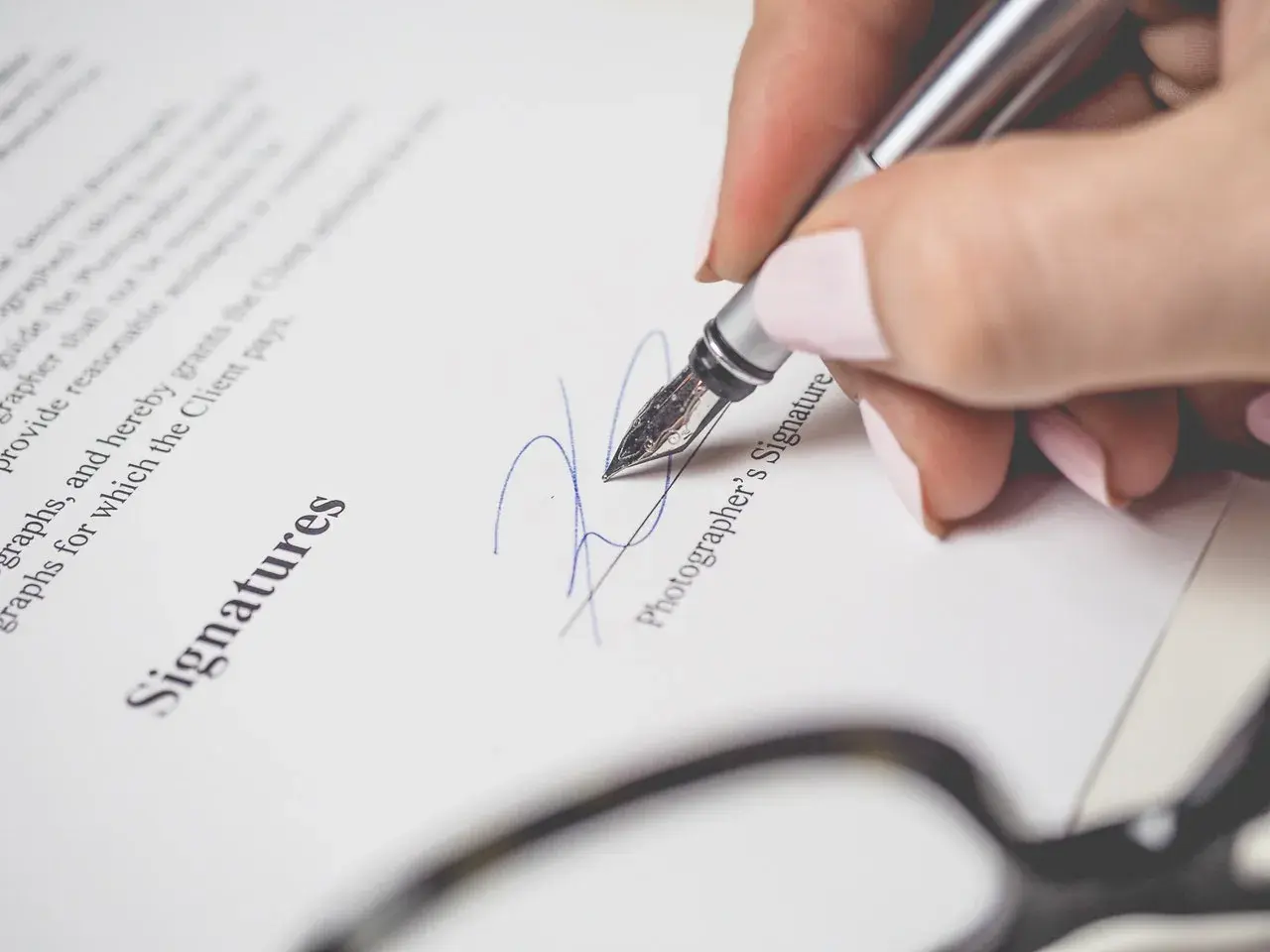 Dłoń z paznokciami malowanymi na różowo podpisuje dokument piórem. Widoczny napis "Signatures" i "Photographer's Signature". To ważny moment, jak wypowiedzenie po angielsku.