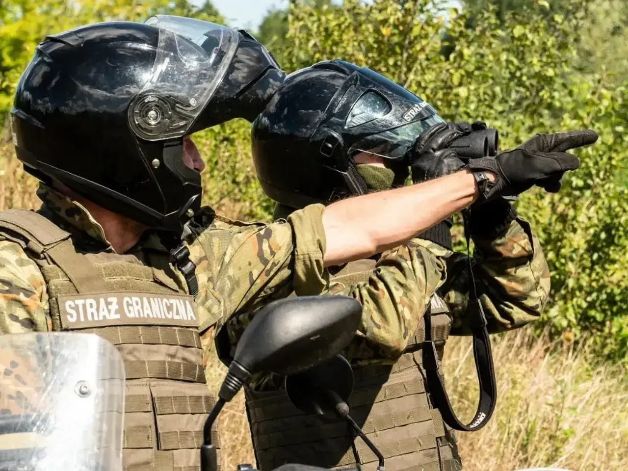 Strażnik graniczny w kamizelce taktycznej i kasku obserwuje teren przez lornetkę, wskazując kierunek.