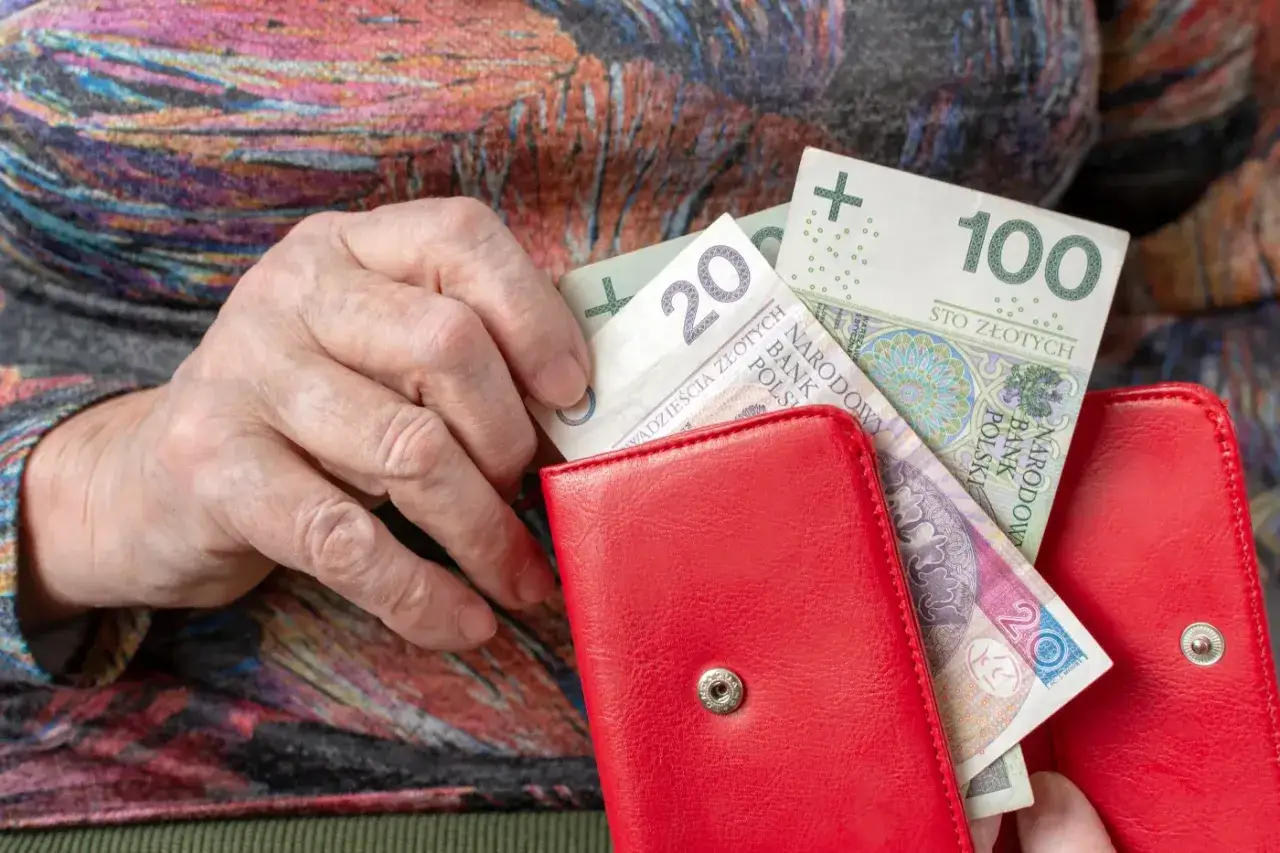 Dłoń starszej osoby wkłada banknoty do czerwonego portfela. To ważne dla najnowszych wiadomości o emeryturach i rentach.
