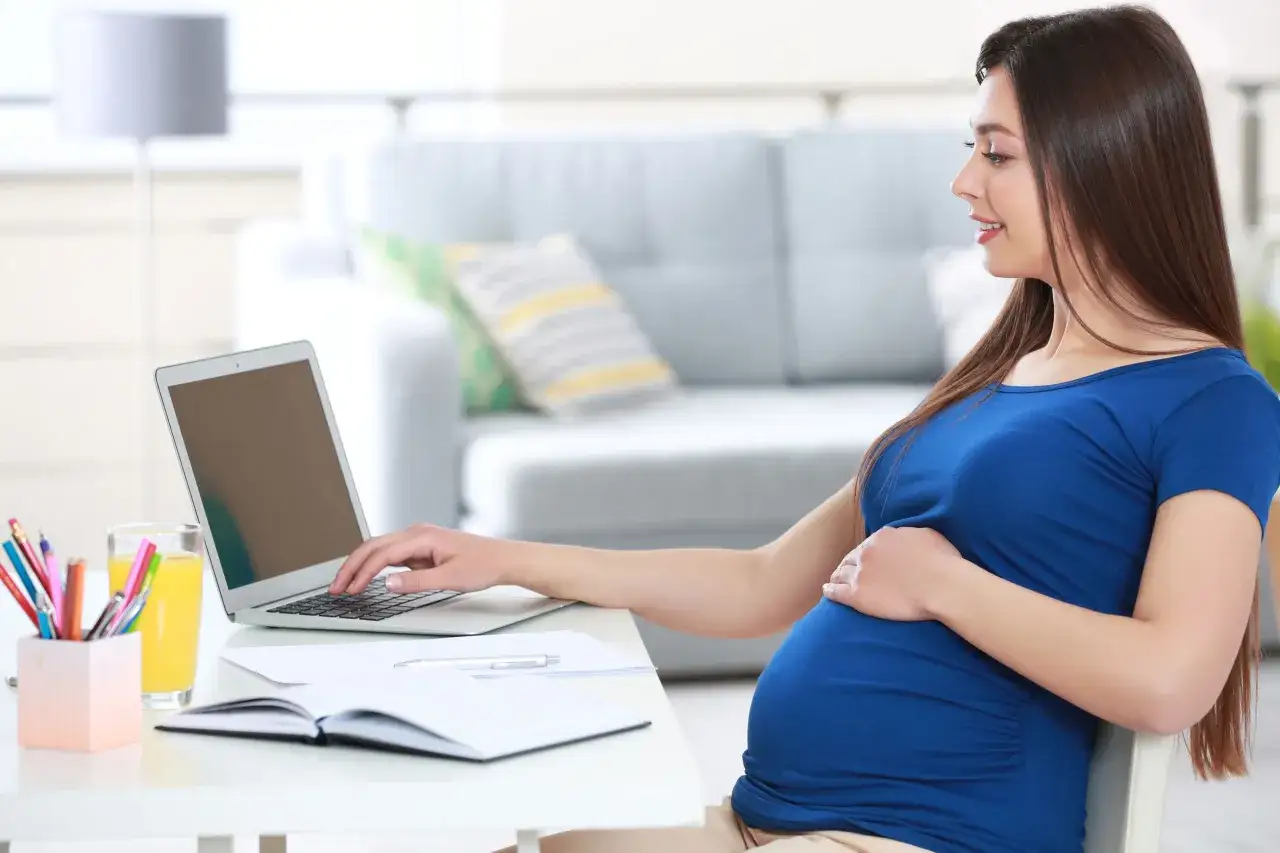 Kobieta w 4 miesiącu ciąży pracuje przy laptopie, opierając dłoń na brzuchu.