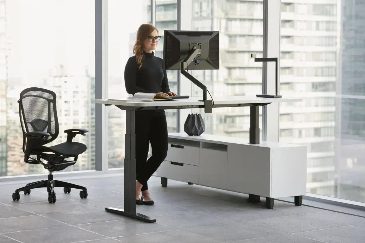 Kobieta pracuje przy biurku z regulacją wysokości, dbając o ergonomię pracy. Obok stoi wygodne krzesło biurowe.