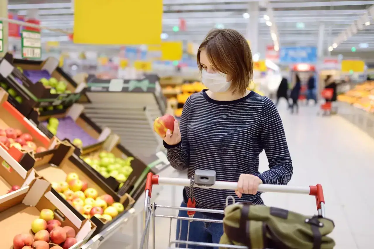 Młoda kobieta w masce wybiera jabłko w supermarkecie. Wózek sklepowy i skrzynki z owocami sugerują, że jest ona klientką, a nie sprzedawcą.
