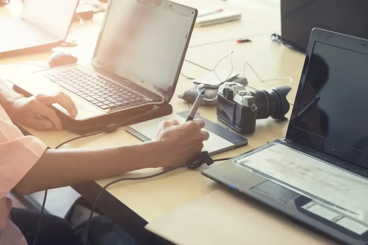 Osoba pracuje na tablecie graficznym i laptopie, obok leży aparat fotograficzny. To idealne warunki do dodatkowej pracy.