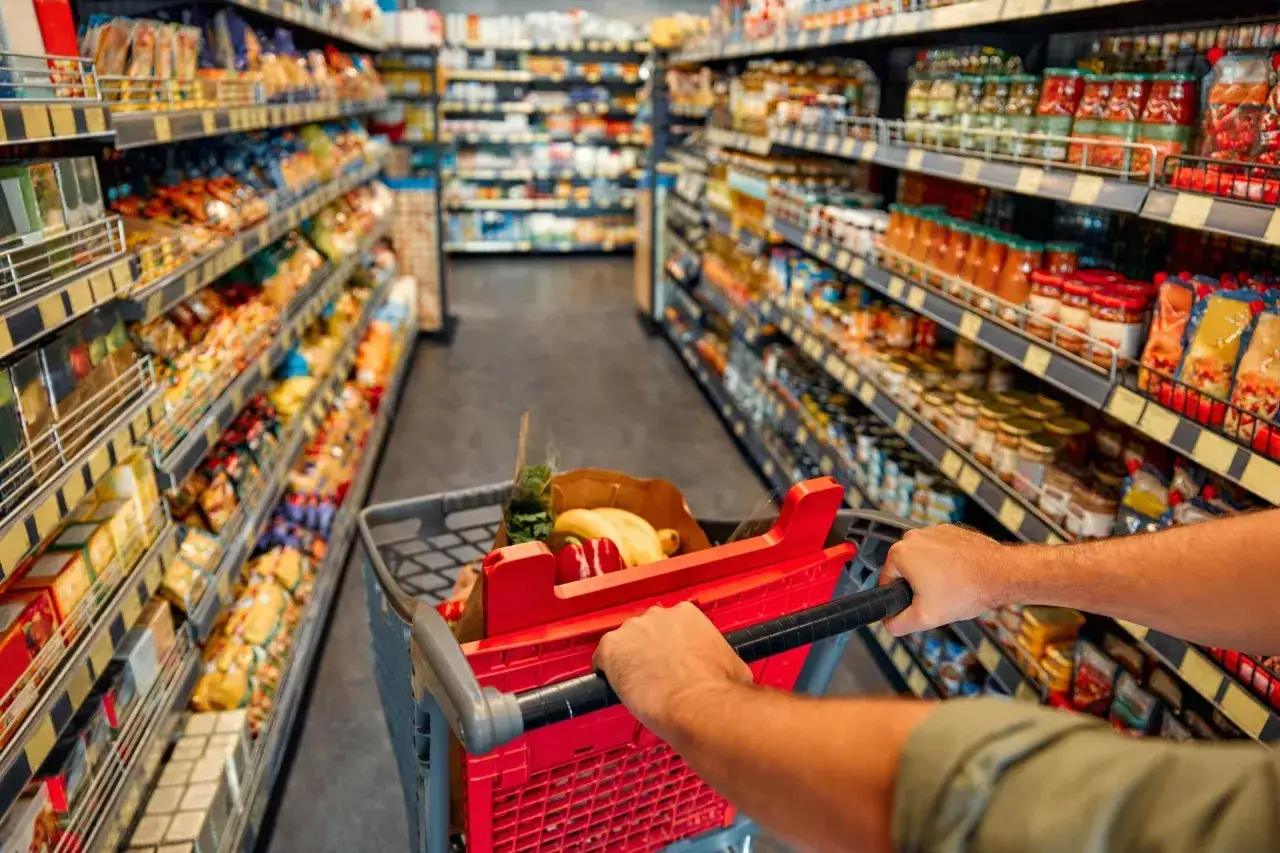 Wózek z zakupami w alejkach sklepu spożywczego. Pełne półki produktów z branży FMCG, od przekąsek po przetwory.